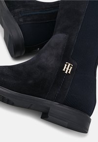 Bottes noires en daim jusqu'aux genoux avec un panneau élastique côtelé, un accent logo en or, une semelle texturée et une fermeture éclair pour un enfilage facile.