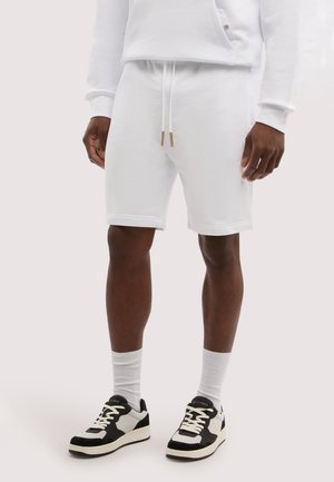 Mężczyzna ubrany w białe szorty ze sznurkiem, białe skarpetki, czarno-białe trampki oraz białą koszulę z długim rękawem, stojący na jednolitym tle.