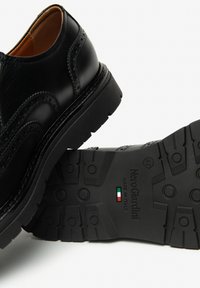 Scarpa elegante in pelle nera con dettagli di brogue decorativi, dotata di una suola in gomma robusta con grip testurizzato e branding italiano sulla base.