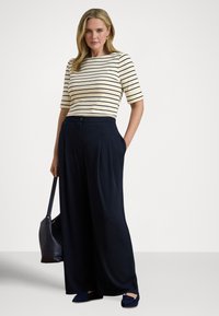 Sieviete, tērpusies krēmkrāsas un melnu svītru topā ar elkoņa garuma piedurknēm, platām melnām biksēm, melnās loafer kurpēs un turot melnu rokassomiņu.