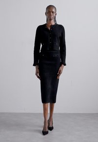 Mustat neulottu kahden osan asukokonaisuus, joka sisältää lyhyen nappitoppi, jossa on taskut, sekä tiukasti istuvan midi-hameen. Asusteena mustat korkokengät.