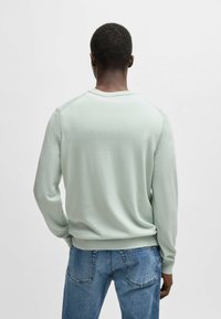Maglione a maglia verde chiaro con scollo rotondo, polsini e orlo a costine; indossato con jeans di denim blu, evidenziando una texture liscia.