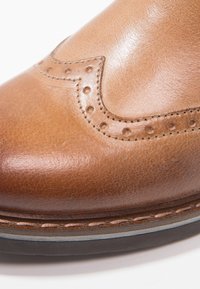 Gros plan sur l'avant d'une chaussure habillée en cuir fauve avec perforations brogue et coutures détaillées sur la vigogne et la tige.