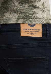 Vihreäkuosisesta paidasta näkyy osia tummien farkkujen päältä. Nahkainen etiketti on ommeltu takataskuun, jossa lukee "Denim Driven Since 1989."