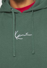 Felpa verde scuro realizzata in tessuto morbido, con logo ricamato bianco. Include lacci e cappuccio foderato. Camicia rossa visibile sotto.