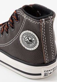 Brązowe skórzane sneakersy za kostkę z pomarańczowymi i czarnymi sznurówkami, białymi przeszyciami oraz okrągłą łatą z logo po boku. Czarna teksturowana obroża.