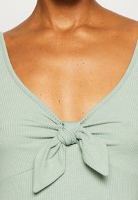 Haut côtelé vert clair avec un profond décolleté en V et un nœud à l'avant, en tissu lisse avec une peau texturée visible en arrière-plan.