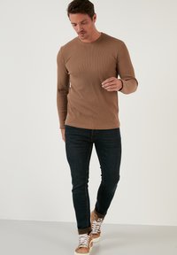 Braun geripptes Langarmshirt kombiniert mit dunkelblauen Jeans und hellbraunen Sneakers, die weiße Schnürsenkel und einen braunen Akzent um den Knöchel haben.