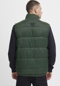 Zeleni puffer prsluk s visokim ovratnikom, koji sadrži kovane panele, teksturiranu površinu i malu oznaku logotipa blizu ovratnika.