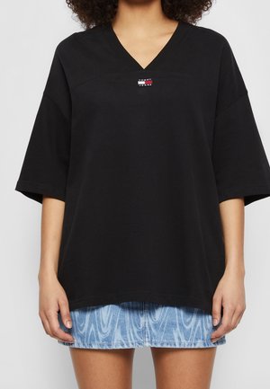 Kobieta w czarnej oversize koszulce Tommy Jeans z dekoltem w serek oraz niebieskiej mini spódnicy jeansowej w wzory, ręce swobodnie opuszczone wzdłuż ciała.