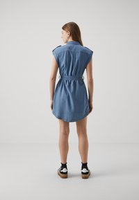 Denim haljina svijetloplave boje, kratki rukavi, strukirana s remenom, zakrivljeni rub i detaljno šivanje. Uparena s crnim čarapama do gležnjeva i tenisicama.