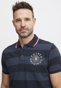 Polo tričko s námorníckym a svetlomodrým pruhovaním, so rebrovým golierom a červenými akcentmi. Na hrudi je vyšívané logo. Krátke rukávy.