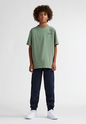 Grünes Baumwoll-T-Shirt mit kurzen Ärmeln und rundem Ausschnitt, kombiniert mit marineblauen Jogginghosen und weißen Sneakern. Einfaches Design ohne Muster.