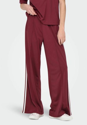 Pantalons larges rouges avec des rayures blanches sur les côtés, fabriqués en tissu lisse. Stylés avec un haut ample et des baskets blanches.