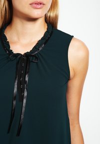 Top sin mangas de color verde oscuro con un escote con volantes, presenta un cierre de lazo y botones, hecho de una tela suave con un ligero brillo.