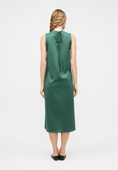 Γυναίκα με μακριά κυματιστά μαλλιά, φορώντας πράσινο midi φόρεμα χωρίς μανίκια με δέσιμο στο λαιμό, στέκεται με την πλάτη προς τα εμπρός σε απλό φόντο.