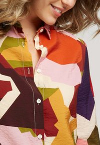 Chemise à boutons colorée présentant un mélange de grandes formes abstraites dans des nuances de rouge, orange, rose, vert et marron. Texture douce avec un col.