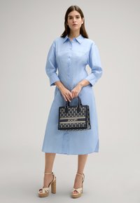 Helles blaues Hemdkleid mit Knopfleiste, langen Ärmeln und Seitenschlitzen, kombiniert mit einer schwarzen und goldfarbenen Handtasche mit Muster, die das Logo "JOOP!" aufweist.