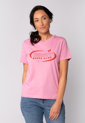 Frau mit dunklem Haar, die ein pinkfarbenes "Beauty Babes Club"-T-Shirt und blaue Jeans trägt, posiert vor einem schlichten hellen Hintergrund.