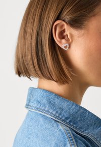 Silberner herzförmiger Steckerohrring mit klaren Verzierungen, am Ohrläppchen befestigt. Das Modell trägt eine blaue Jeansjacke, die Haare leicht über schulterlang.