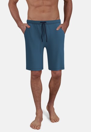 Mann trägt blaue knielange Shorts mit Kordelzug und Taschen, steht barfuß vor einem schlichten hellen Hintergrund.