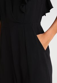 Monos de color negro con un escote con volantes y mangas cortas, con un bolsillo lateral y un corte entallado, fabricado en tejido suave.