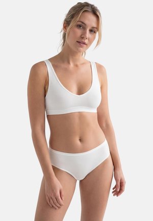 Frau mit blondem Haar, die einen weißen BH aus weichem Stoff und passende weiße Slips trägt, vor einem schlichten hellen Hintergrund steht.