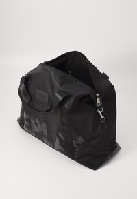 Borsa tote in tessuto nero con finitura opaca, dotata di chiusura con zip, manici piatti e logo in rilievo. Grande dimensione con design minimalista.