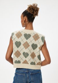 Strikket ærmeløs sweater med cremefarvet baggrund, prydet med grønne hjerter og brune diamantmønstre. Ribbet halsudskæring og cropped design.