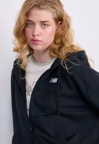 Svart zip-up hettegenser med kengurulomme og snorhette. Har en hvit New Balance-logo på brystet. Mykt, glatt stoff.