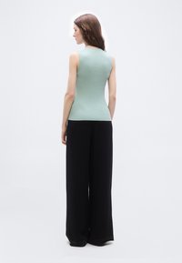 Γυναίκα με μακριά καστανά μαλλιά που φοράει αμάνικο στενό ανοιχτό πράσινο τοπ και φαρδιά μαύρη παντελόνα, όρθια με την πλάτη γυρισμένη σε λευκό φόντο.