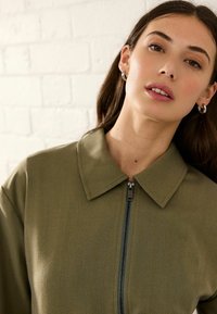 Olivgrüne Jacke mit Fischgrätenstruktur, die einen Kragen und einen Reißverschluss auf der Vorderseite hat.
