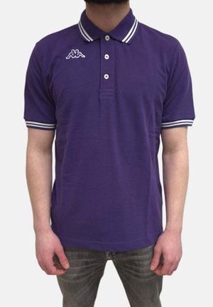 Uomo con polo viola a maniche corte con colletto e maniche a righe bianche, in piedi davanti a uno sfondo semplice.