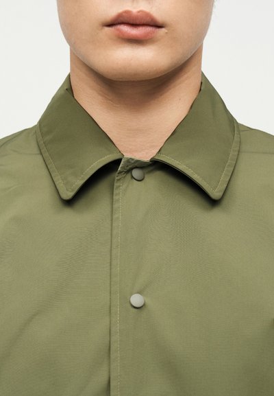 Κοντινό πλάνο του κάτω μέρους προσώπου και του λαιμού ενός ατόμου που φοράει μια πουκάμισο ελιάς με γιακά, κουμπωμένη μέχρι το επάνω κουμπί.