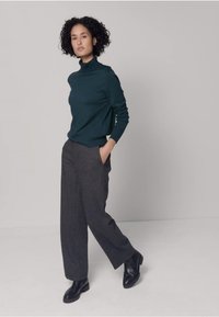 Pull à col roulé vert foncé en tissu lisse, associé à un pantalon gris à jambes larges. Le mannequin pose avec assurance, les mains dans les poches, portant des bottes noires.