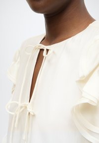 Λευκό μπλουζάκι με βαθύ V-λαιμό, κλείσιμο με κορδόνι και μανίκια με βολάν. Το ύφασμα φαίνεται ελαφρύ με λεία υφή.