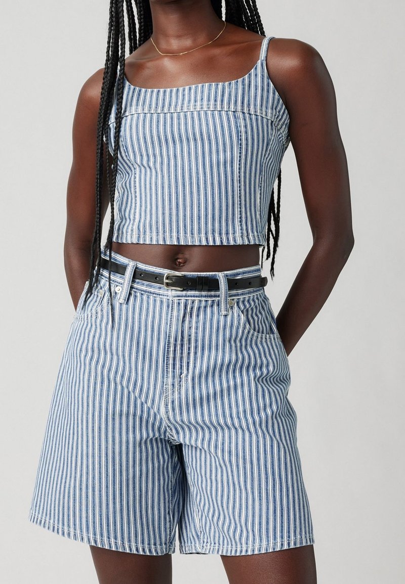 Jeune personne à la peau foncée portant un crop top sans manches à rayures verticales bleues et blanches et un short taille haute assorti avec une ceinture noire.