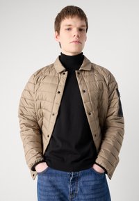 Veste matelassée beige avec fermeture à boutons-pression, poignets côtelés et poche latérale ornée de détails en métal noir, portée sur un col roulé noir.