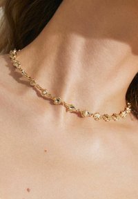 Gylden choker-halskæde med uregelmæssige, strukturerede led og indlagte klare ædelsten, båret på en bar hals. Blød hudfarvet baggrund.