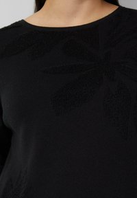 Frau trägt einen schwarzen Strickpullover mit strukturierten Blumenmustern in der Nähe des Halsausschnitts und des Schulterbereichs.