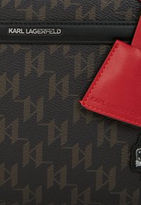 Geantă neagră cu textură, cu un model geometric maro, având un tag din piele roșie și un accent de logo argintiu. Branding-ul distinctiv al designerului este vizibil.