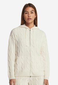 Krämfärgad kabelstickad hoodie med dragkedja fram, huva med dragsko och ribbade ärmslut. Innehåller sidfickor och ett strukturerat mönster genom hela plagget.