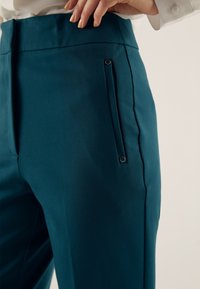 Pantalon bleu sarcelle avec une ceinture plate, une poche latérale ornée d'un petit détail en métal, et une texture de tissu lisse. Coupe cintrée, design minimaliste.