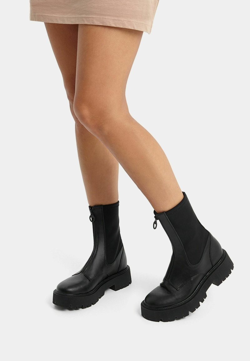 Jambes portant des bottines noires à semelle épaisse avec zips devant et panneaux latéraux élastiques, associées à une jupe beige.