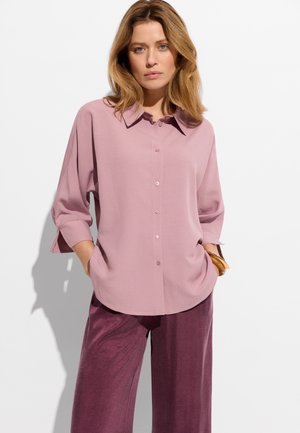 Kobieta o blond włosach, ubrana w luźną, blado-różową koszulę na guziki i szerokie, fiołkowe spodnie, stojąca z rękami w kieszeniach.