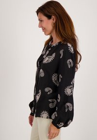 Zwarte blouse met lange mouwen, voorzien van een wit paisleypatroon, met knopen aan de voorkant en licht opbollende mouwen, gemaakt van een lichtgewicht stof.