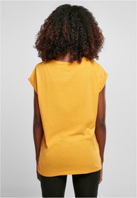 Keltainen puuvillapaita, jossa on lyhyet hihat, rento istuvuus ja pyöreä kaula-aukko, kuvattuna takaa yksinkertaista taustaa vasten.