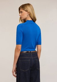 Tricou polo albastru, ajustat, din material tricotat, cu mâneci scurte și un guler moale, asortat cu pantaloni din denim închis la culoare, cu accente de buline aurii.