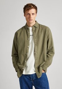 Olijfgroen, langarm overhemd met knopen van een gestructureerde stof en witte knopen, gedragen over een wit T-shirt, gecombineerd met blauwe spijkerbroeken.