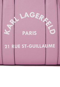 Roze leren tas met geperst witte tekst: "KARL LAGERFELD," "PARIJS," en "21 RUE ST-GUILLAUME," met een gestructureerde rechthoekige vorm en een zachte textuur.